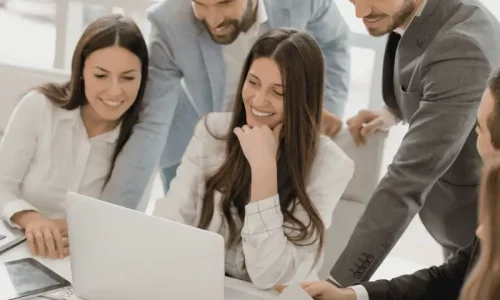 Team of professionals engaged in a collaborative group coaching workshop session, smiling and discussing leadership strategies around a laptop in a modern office setting